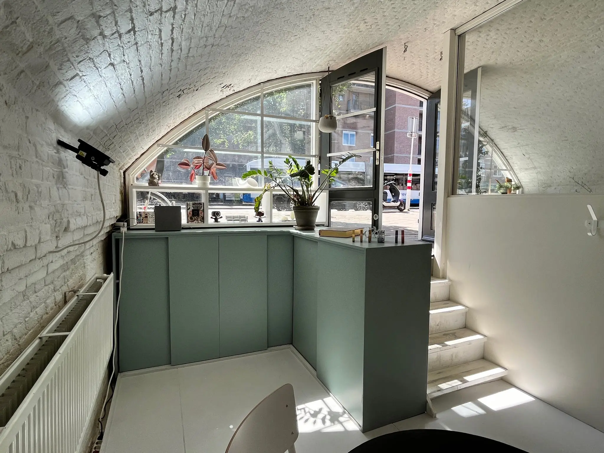 Een modern interieur met gebogen plafond en glazen deur die leidt naar een zonovergoten straat, getiteld 'Tussen de Bogen'.
